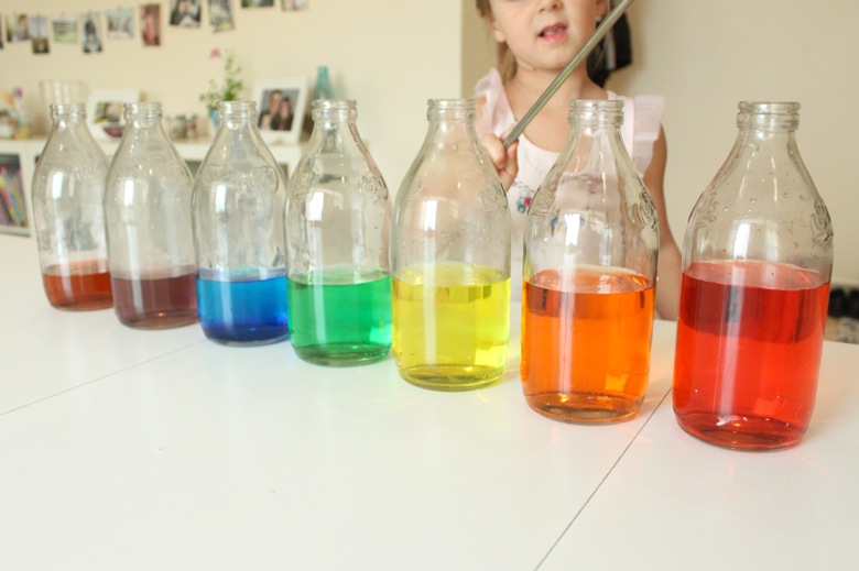 rainbow-xylophone child standing behind row of glass jars with colored water in them