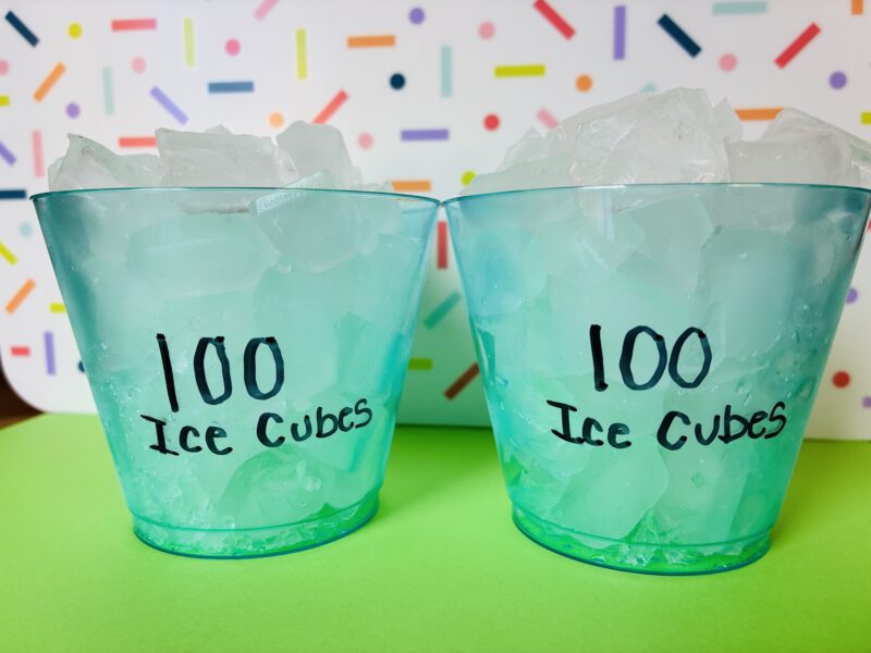 two blue cups with 100 mini ice cubes sitting in front of a rainbow background to conduct a melting ice cube experiment in the class to celebrate the 100th day of school